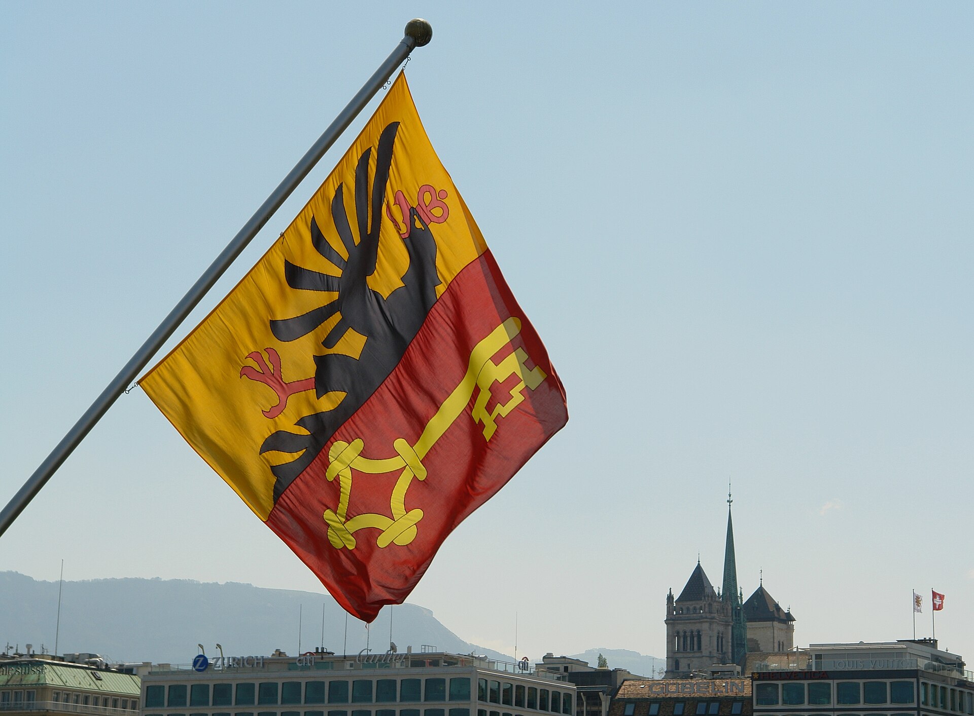 The red and yellow flag of Geneva with its eagle and key flying from a pole, with the towers and spire of St Pierre Cathedral on the skyline behind