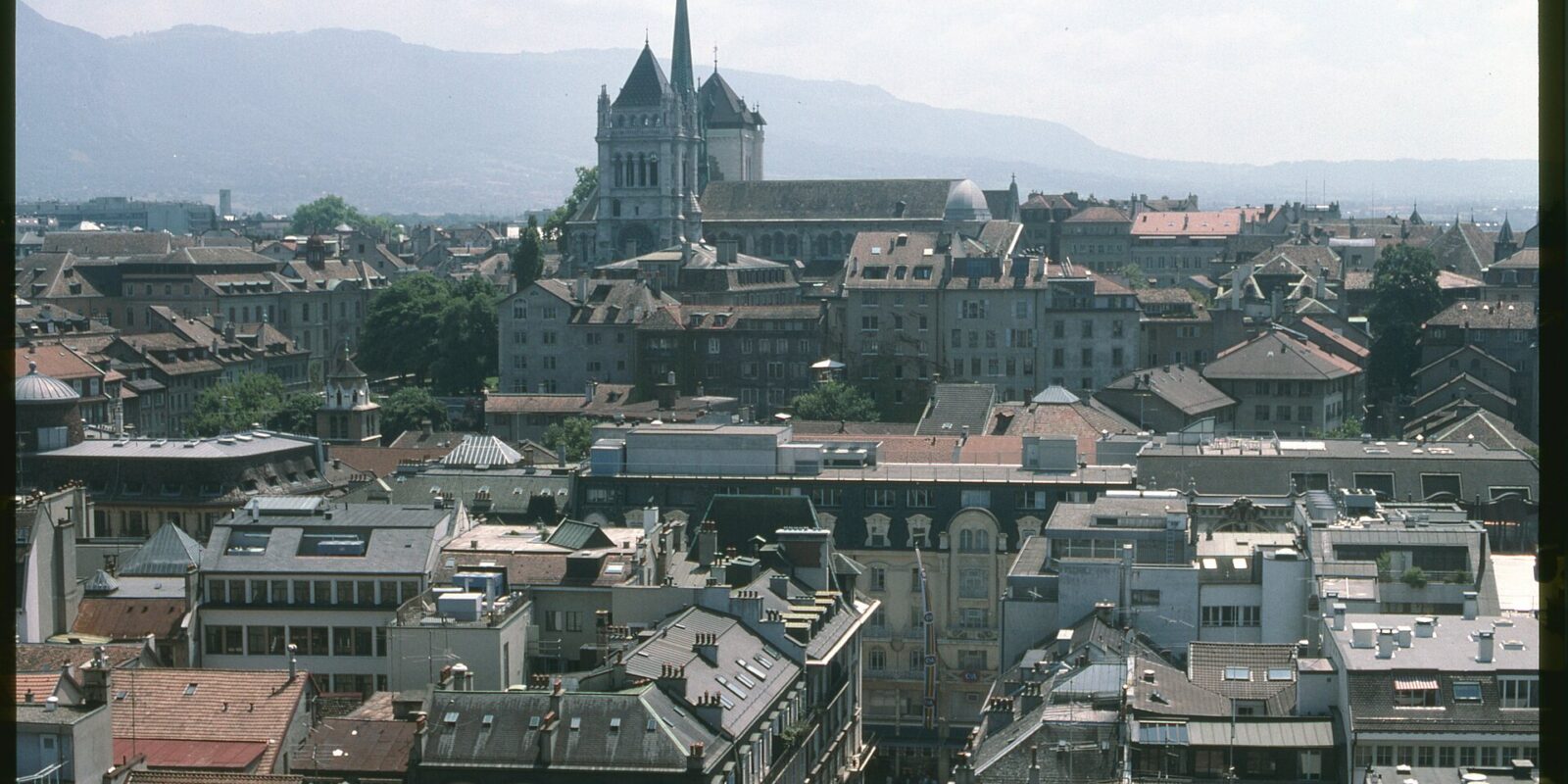 St Pierre Cathedral in Geneva with its towers and green spire rising above the rooftops of the Old Town, with mountains on the horizon