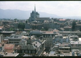 St Pierre Cathedral in Geneva with its towers and green spire rising above the rooftops of the Old Town, with mountains on the horizon