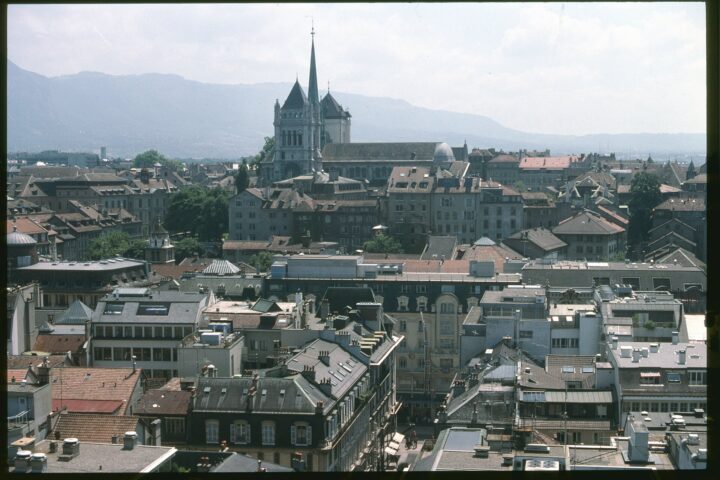 St Pierre Cathedral in Geneva with its towers and green spire rising above the rooftops of the Old Town, with mountains on the horizon
