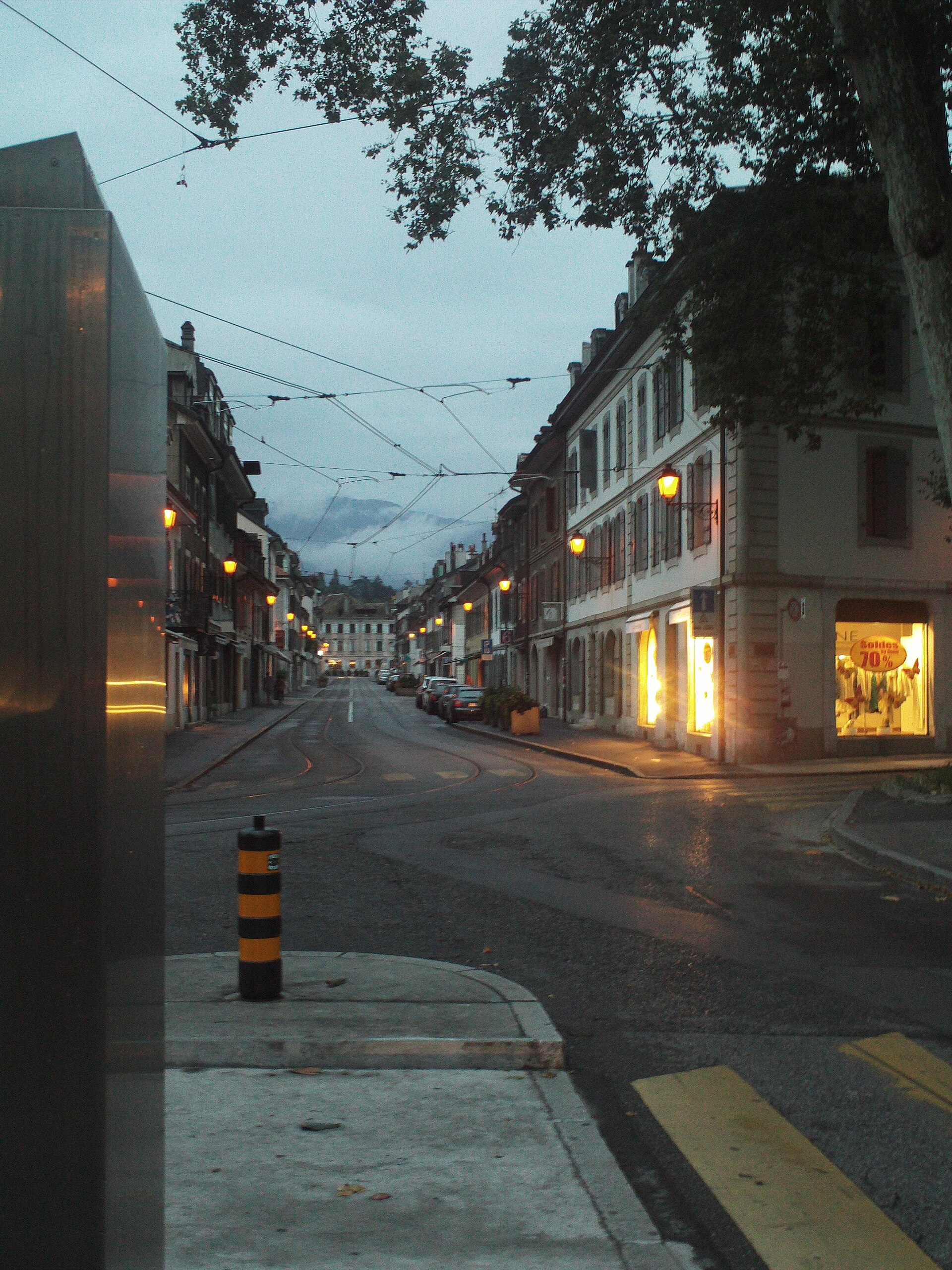 Street lamps and shop windows glow along Rue Caroline in Carouge, where tram tracks curve between low shuttered buildings at dusk.