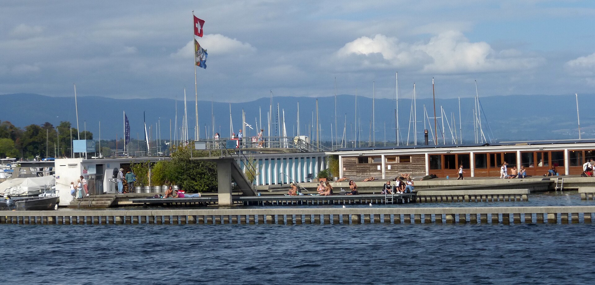 Sunbathers sit and recline beside the water at Geneva’s Bains des Pâquis, with flags and marina masts behind.
