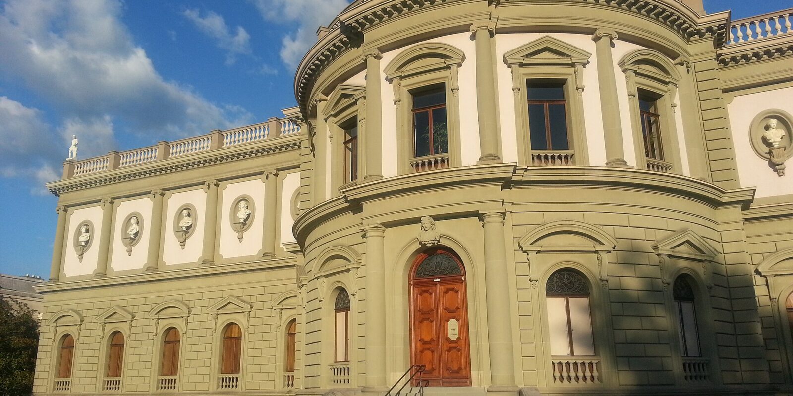 The sunlit Ariana museum in Geneva has a domed central bay, portrait roundels and steps leading to a wooden door.