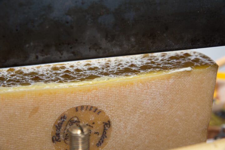 Swiss raclette cheese bubbles beneath a heater, with the rind and stamped Swiss mark visible on the half-wheel.