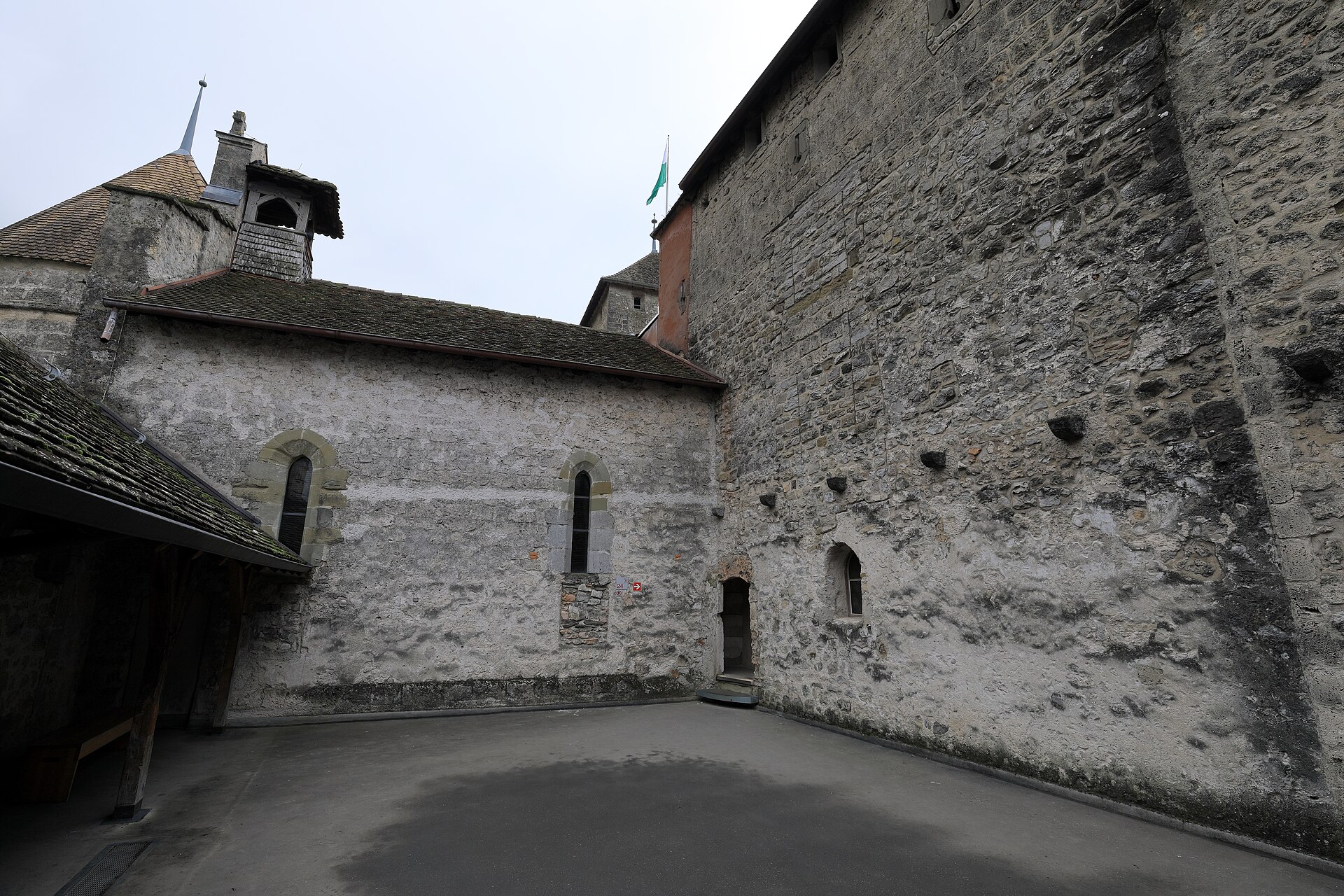 Tall stone walls and narrow windows enclose a quiet courtyard inside Chillon Castle, beside a roofed wooden passage.