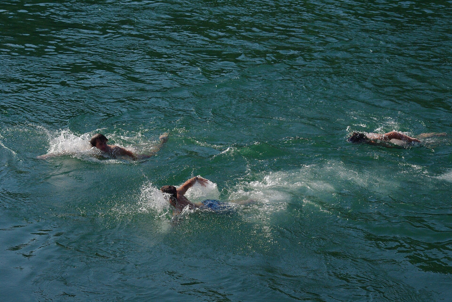 Three swimmers cross the green water of the Rhône at Pont Sous-Terre in Geneva, seen from above.