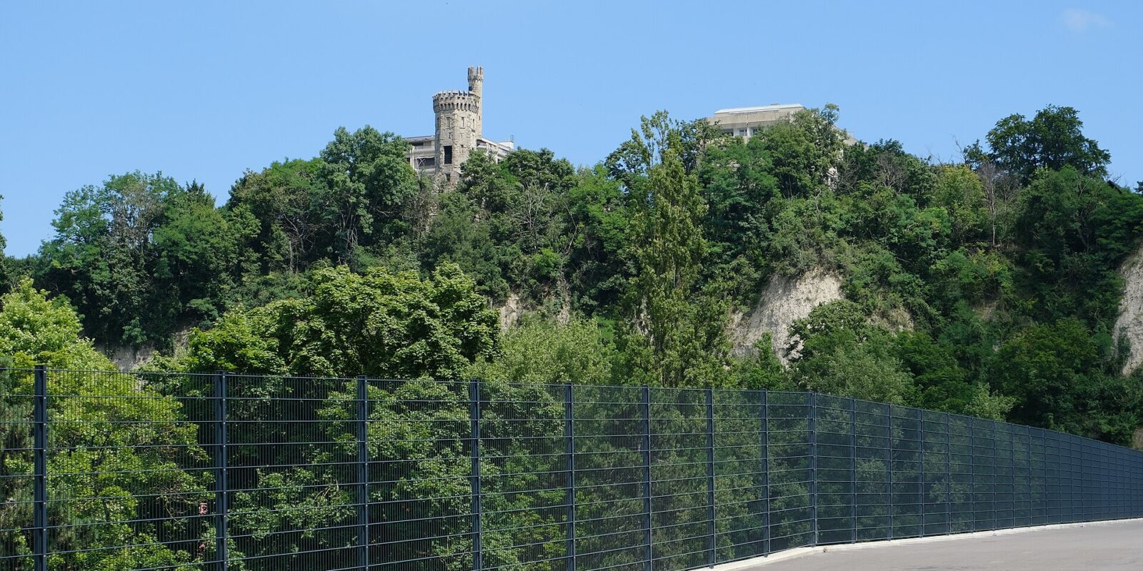 The Tour de Champel rises above wooded Geneva cliffs, viewed from the fenced upper walkway of the Pont des Artisanes.