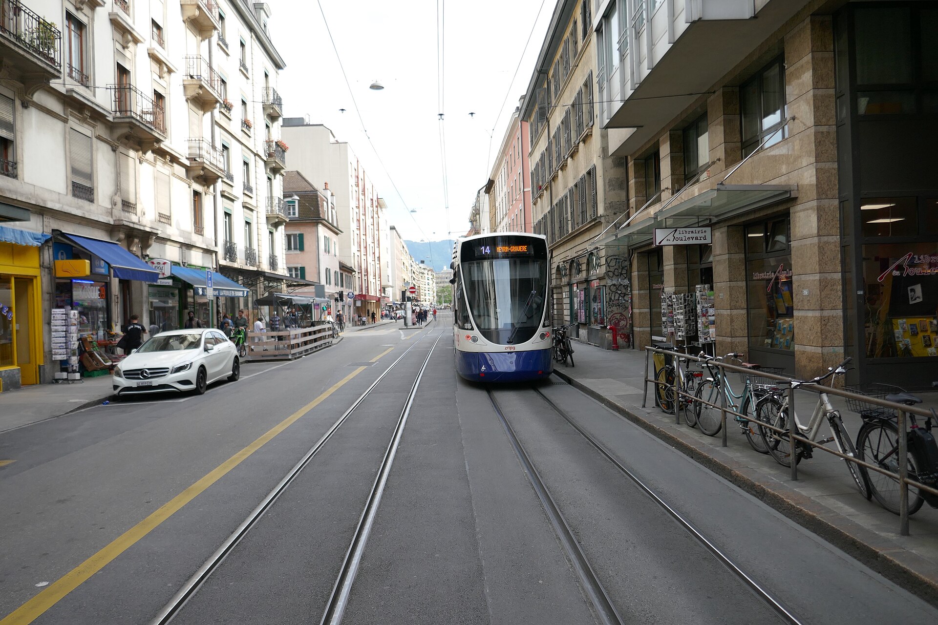 A tram passes shops and a pavement cafe terrace on Rue de Carouge in Geneva's Plainpalais district, with bicycles parked opposite.