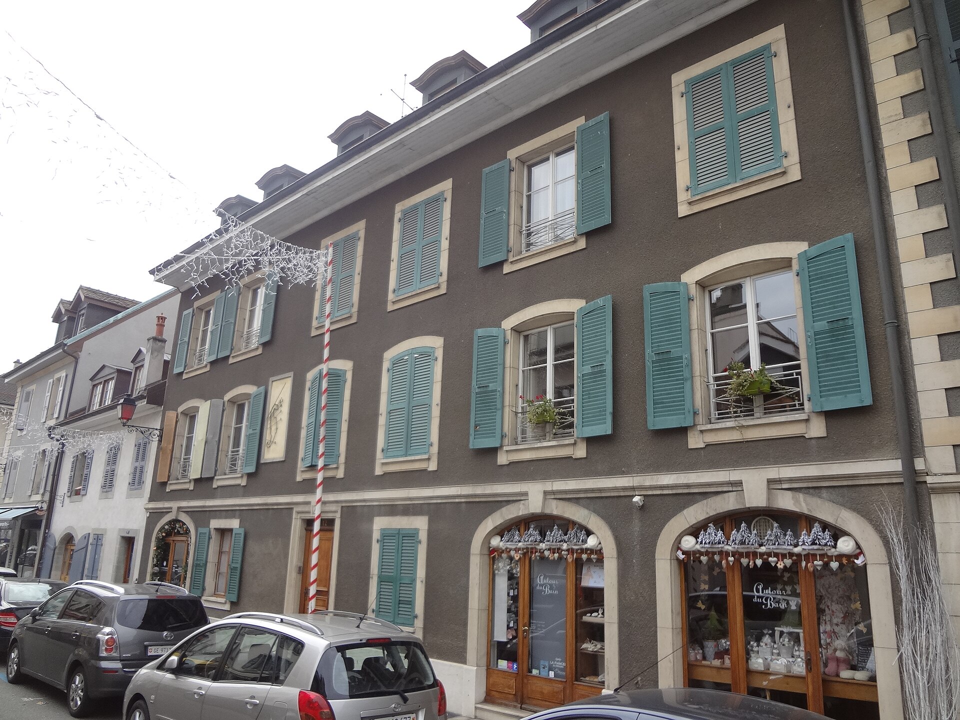 Turquoise shutters and arched shop windows decorate a low grey house on Rue Saint-Joseph in Carouge, with cars parked outside.