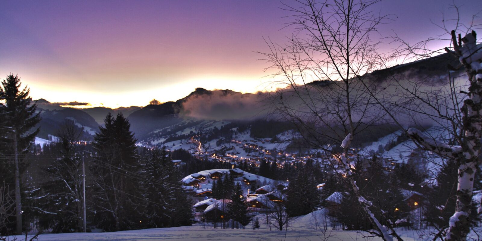 Village lights glow among snow-covered chalets in Les Gets beneath a purple winter twilight sky and surrounding mountain slopes.