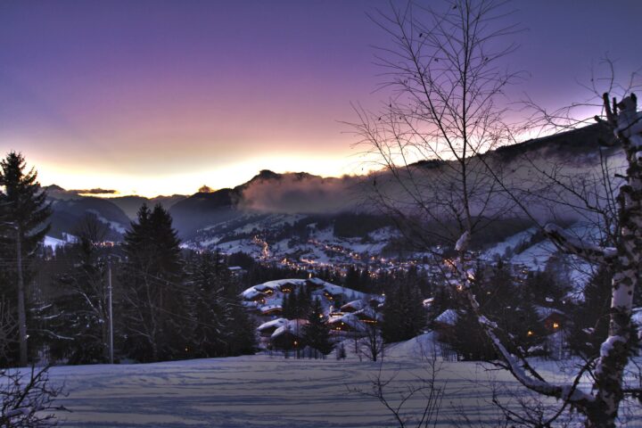 Village lights glow among snow-covered chalets in Les Gets beneath a purple winter twilight sky and surrounding mountain slopes.