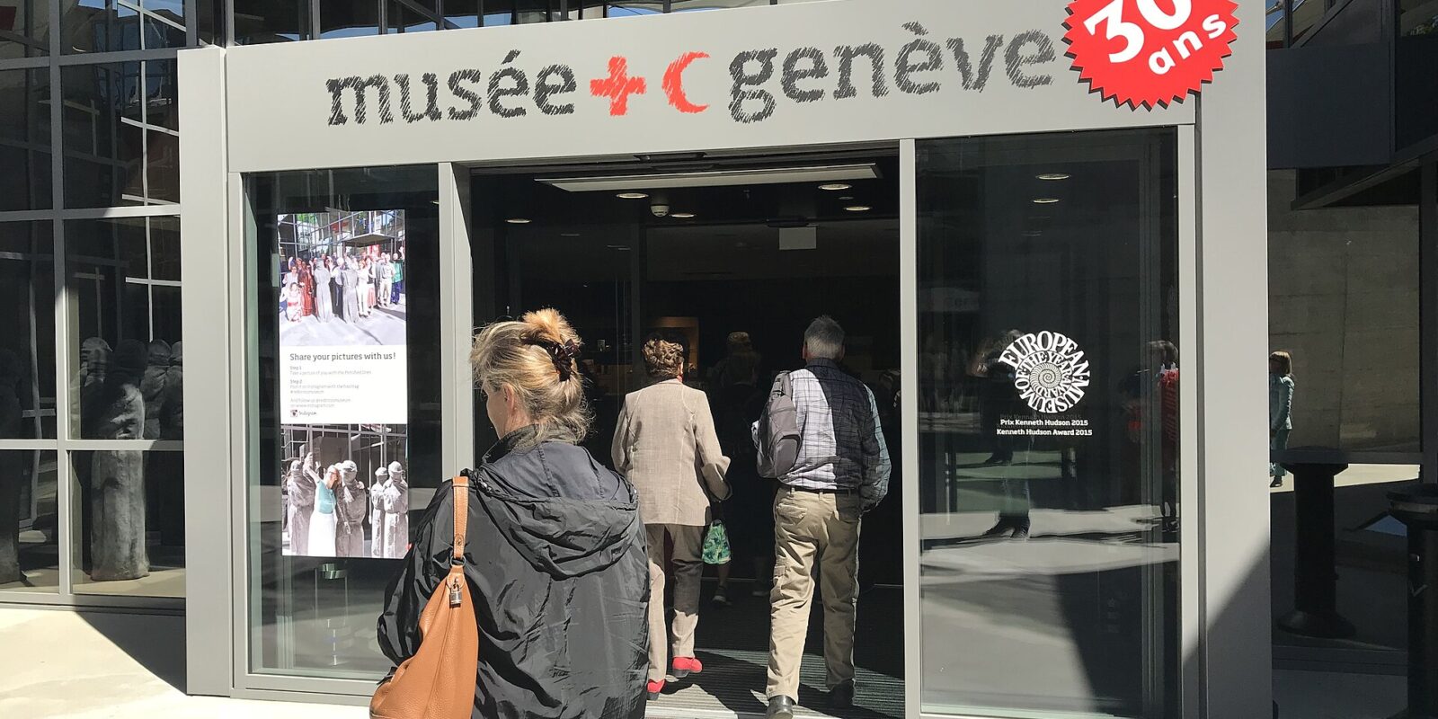 Visitors enter Geneva's International Red Cross and Red Crescent Museum beneath its name and red emblems on the glass entrance.