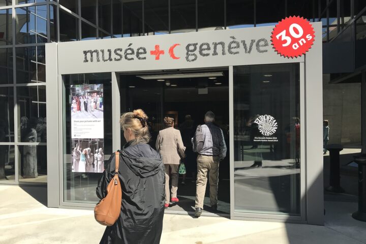 Visitors enter Geneva's International Red Cross and Red Crescent Museum beneath its name and red emblems on the glass entrance.