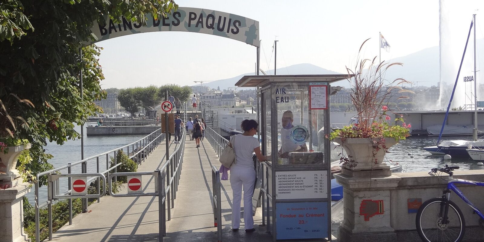 Visitors follow the railed entrance ramp at Bains des Pâquis in Geneva, with the Jet d'Eau rising across the water.