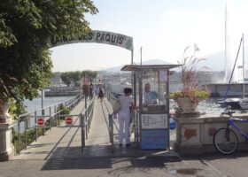 Visitors follow the railed entrance ramp at Bains des Pâquis in Geneva, with the Jet d'Eau rising across the water.