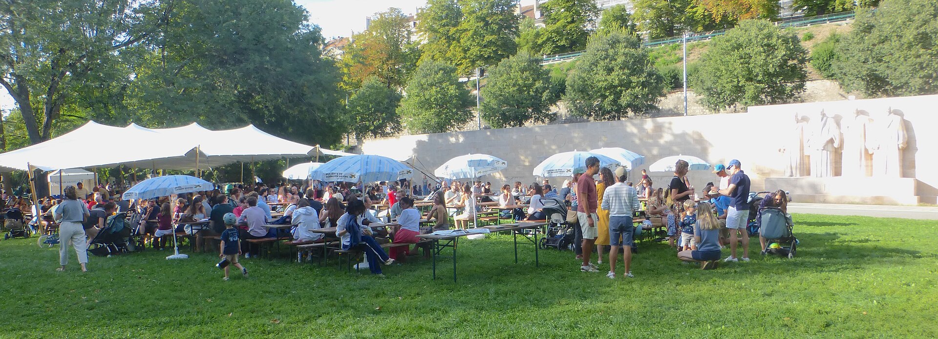 Visitors gather around picnic tables and tents beside the Reformation Wall in Geneva's Parc des Bastions.