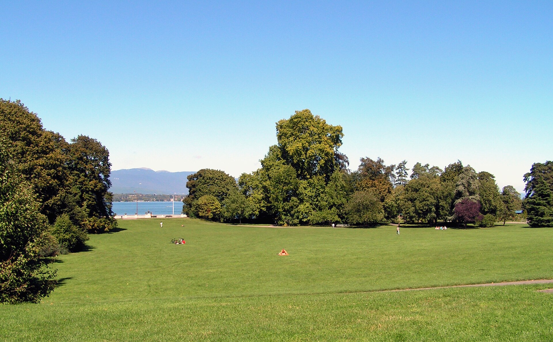 Visitors relax on the broad lawn at Parc La Grange in Geneva, with mature trees and Lake Geneva beyond.