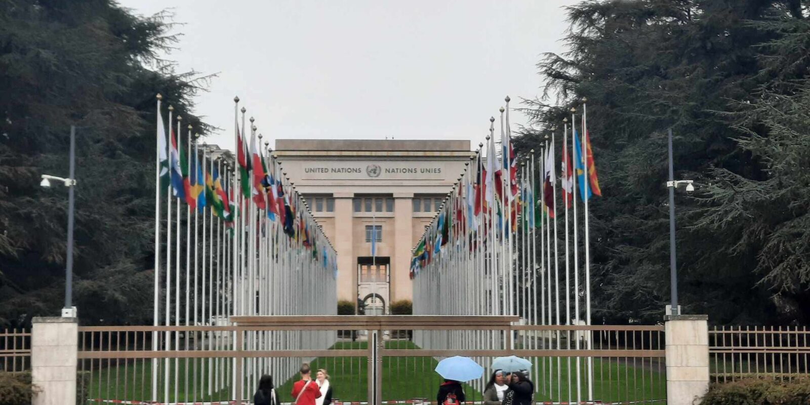 Visitors shelter under umbrellas outside the Palais des Nations in Geneva, with flags lining the approach behind the gates.