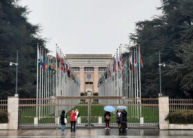 Visitors shelter under umbrellas outside the Palais des Nations in Geneva, with flags lining the approach behind the gates.