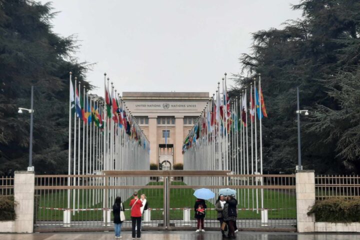 Visitors shelter under umbrellas outside the Palais des Nations in Geneva, with flags lining the approach behind the gates.