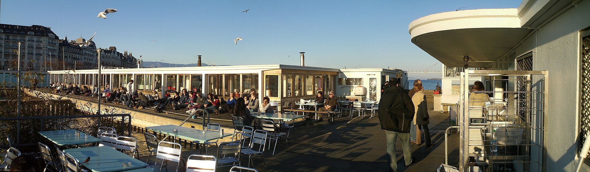 Visitors sit beside the Buvette des Bains terrace in Geneva, with outdoor tables, white baths buildings and gulls overhead.