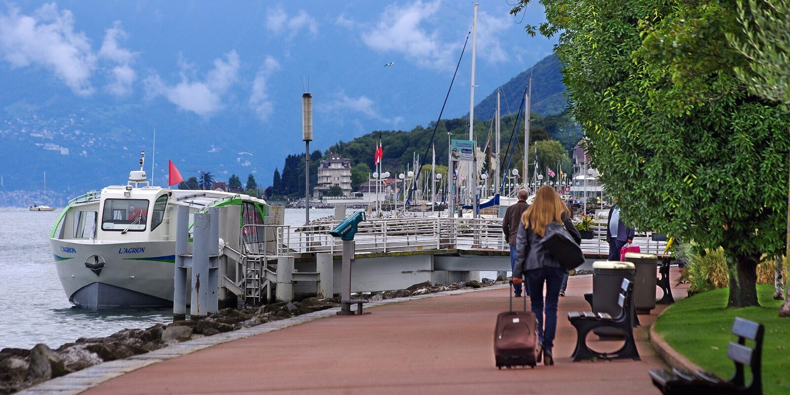 Visitors walk along the tree-lined promenade in Évian-les-Bains beside the moored L'Agrion boat and Lake Geneva.