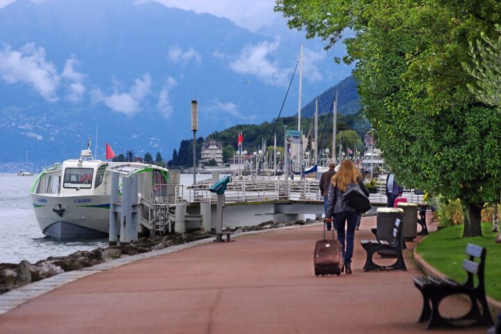 Visitors walk along the tree-lined promenade in Évian-les-Bains beside the moored L'Agrion boat and Lake Geneva.