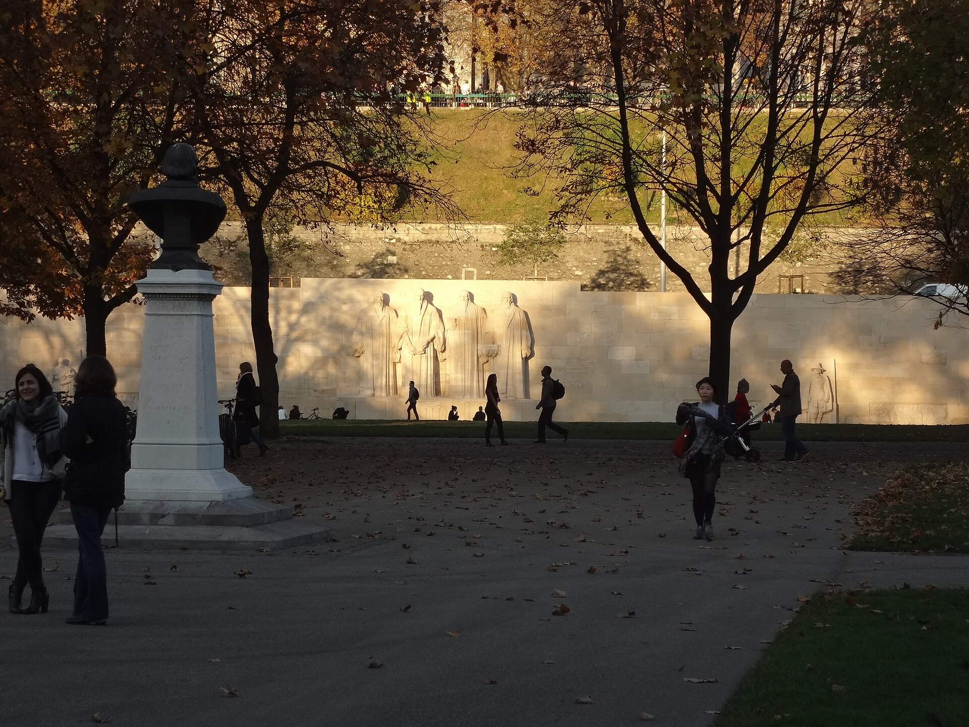 Visitors walk beneath autumn trees in Parc des Bastions, with Geneva's Reformation Wall illuminated beyond them.