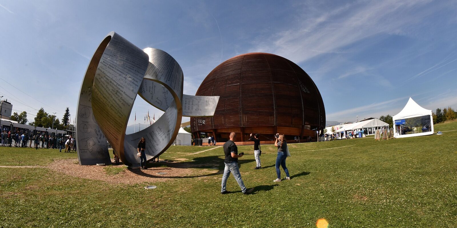 Visitors walk past a curved metal sculpture on the lawn outside the wooden CERN Globe during the 2019 open days.