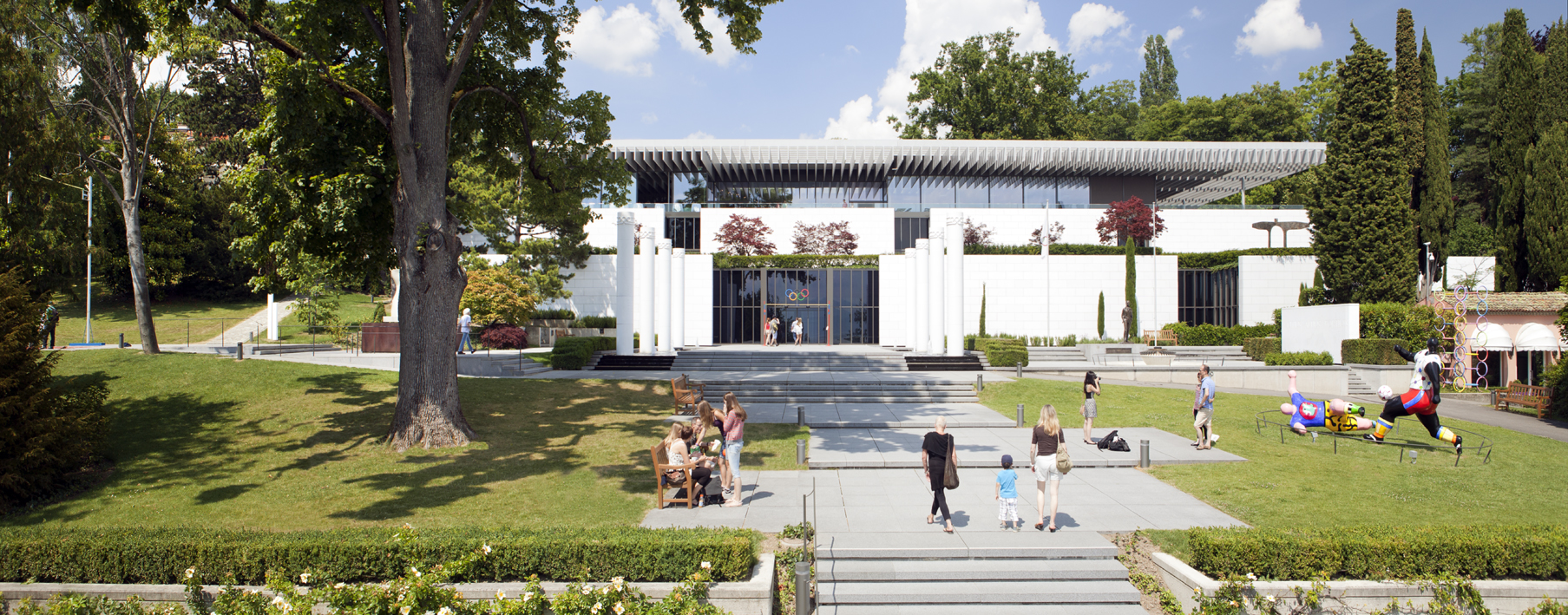Visitors walk through landscaped grounds toward the white Olympic Museum building in Lausanne, with Olympic rings above its entrance.