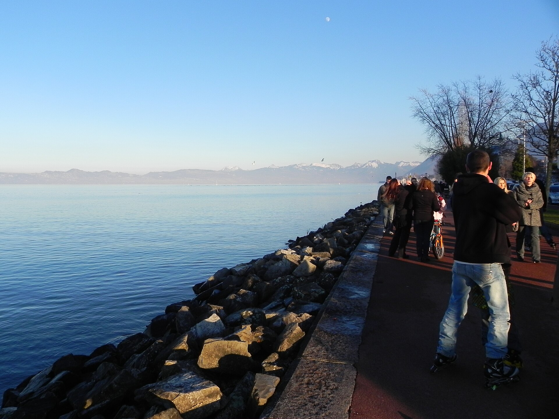 Walkers and a roller skater share Evian’s lakeside promenade beside calm Lake Geneva, with snowy mountains in the distance.