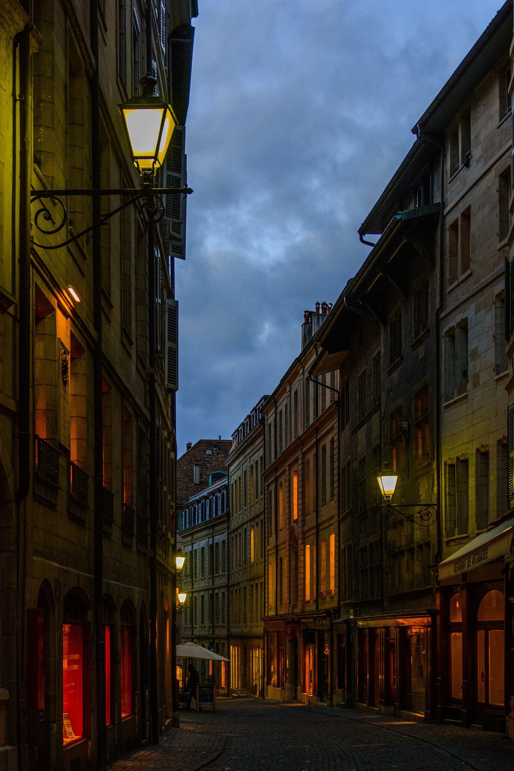 Warm lanterns illuminate the cobbled Grand-Rue between tall stone houses in Geneva Old Town at dusk.