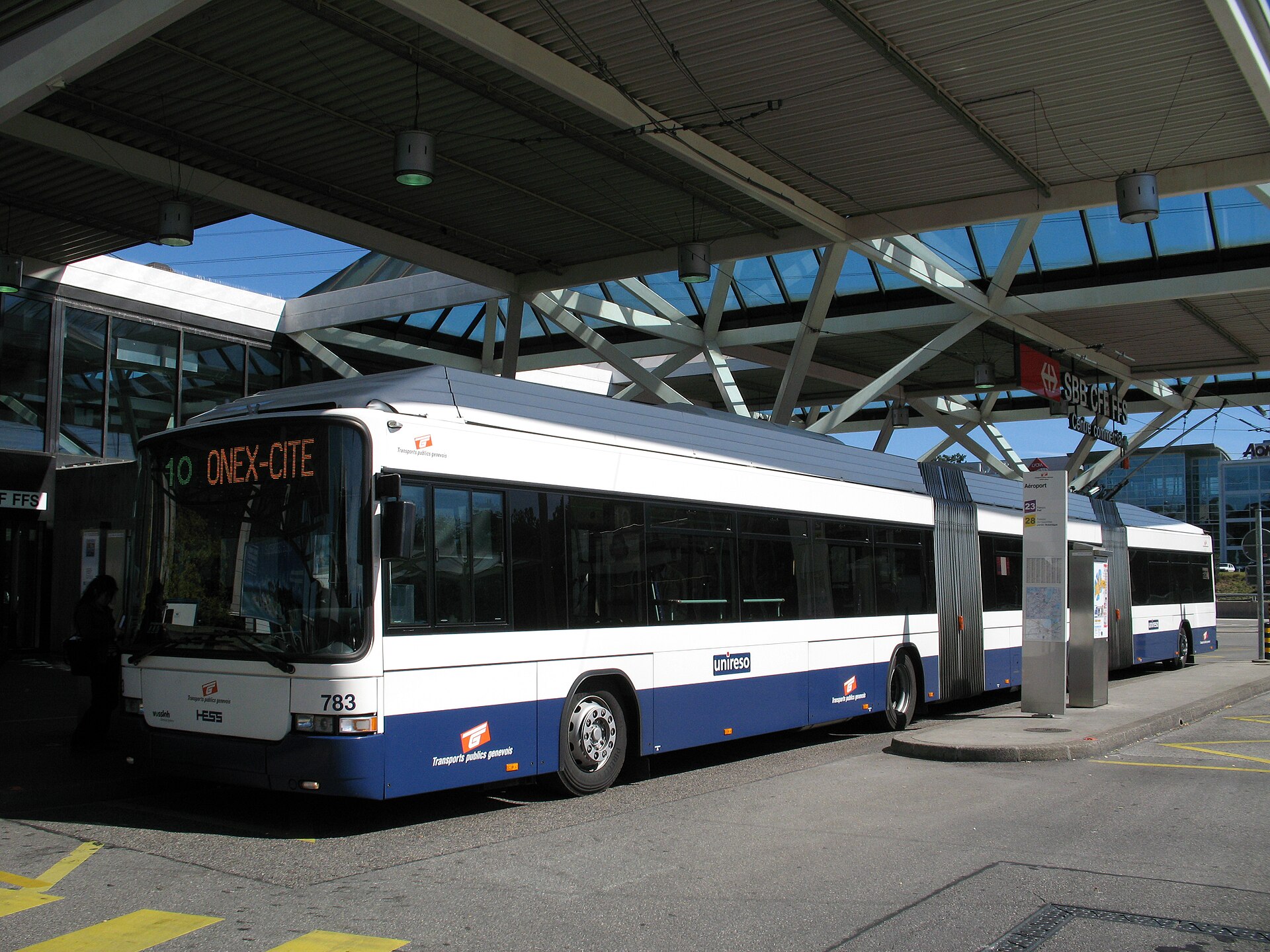 A white and blue articulated TPG trolleybus waits beneath the canopy at Geneva Airport, beside an Aeroport stop sign.