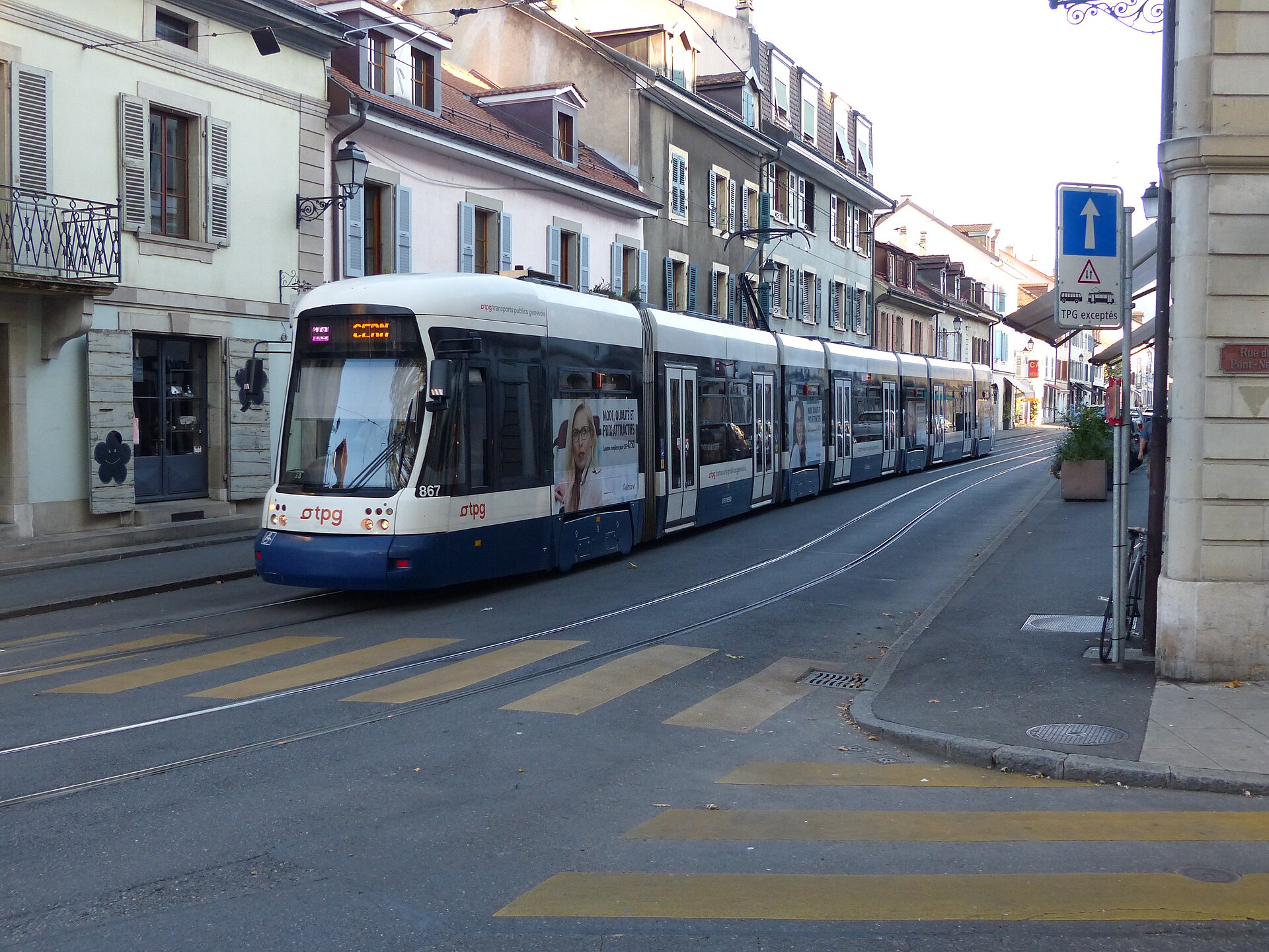 A white and blue TPG tram passes shuttered houses in Carouge beside a yellow pedestrian crossing and overhead wires.