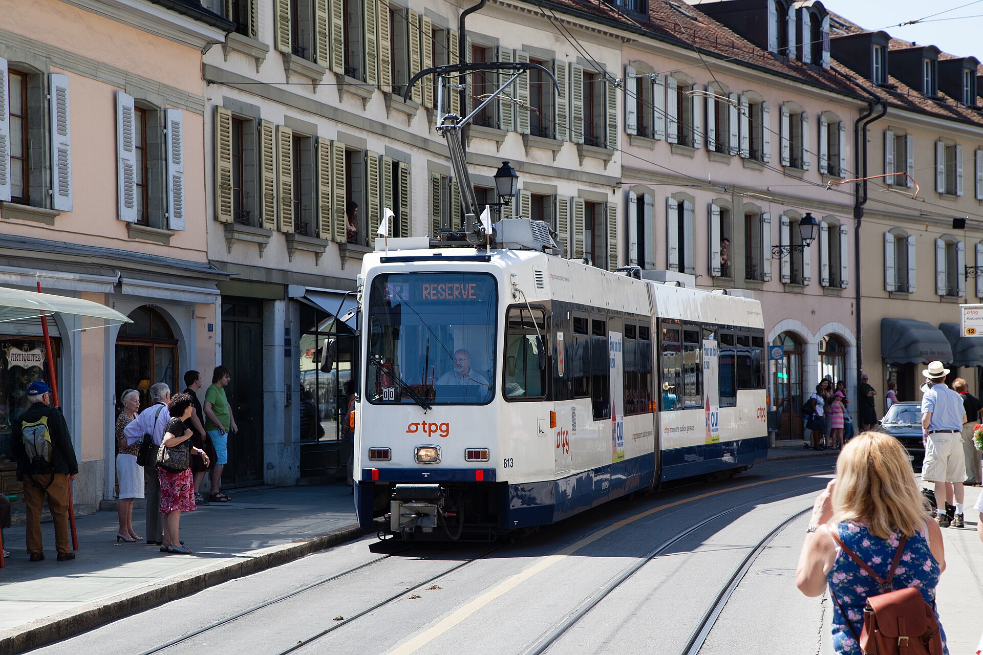 A white and blue TPG tram stands on Rue Ancienne in Carouge beside pedestrians and a row of pale shuttered houses.