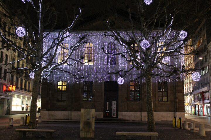 White Christmas lights cover a Geneva church facade, with glowing spheres hanging between bare trees on the square.
