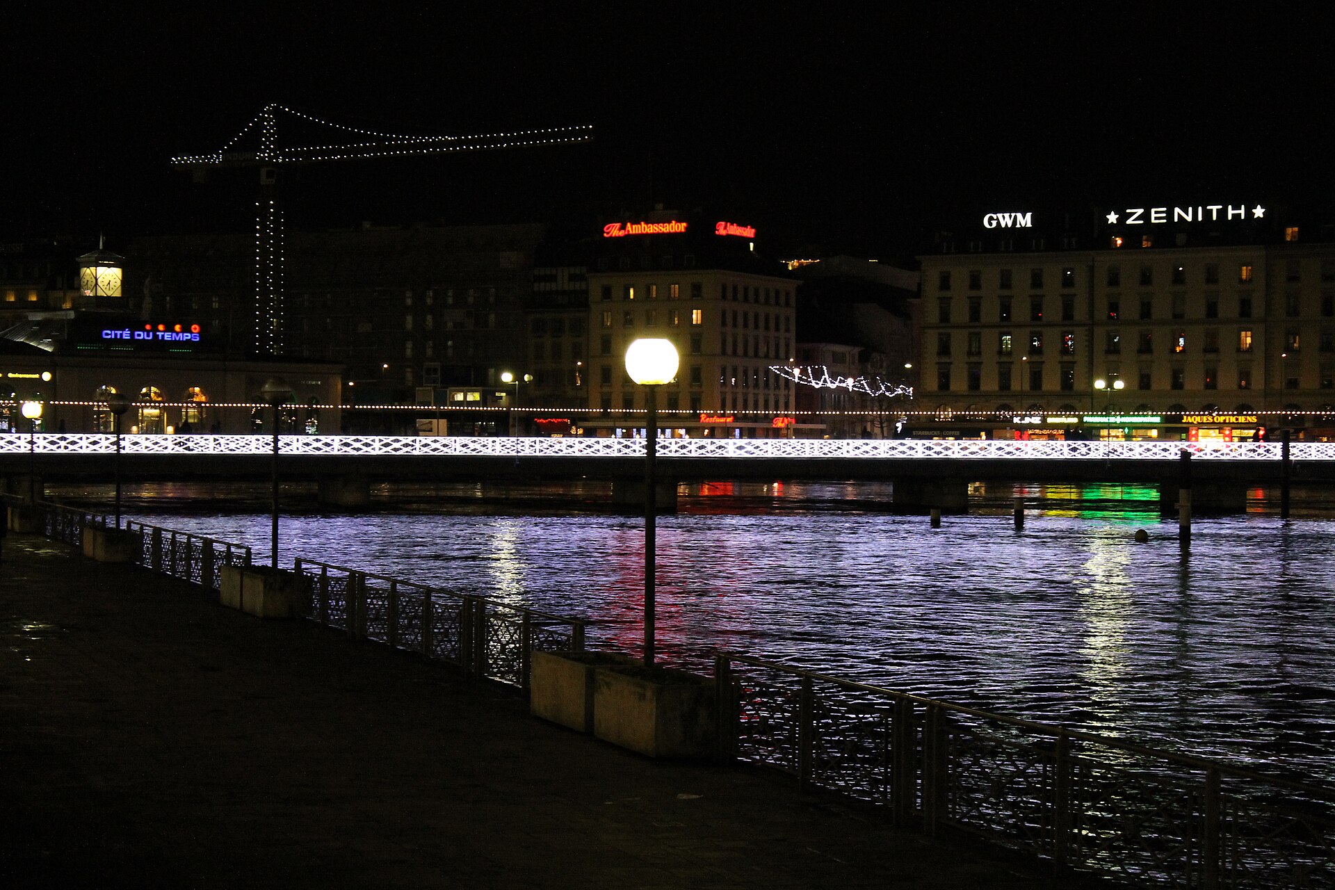 White festive lights and glowing signs reflect in Geneva's waterfront beside a lamp-lit promenade after dark.