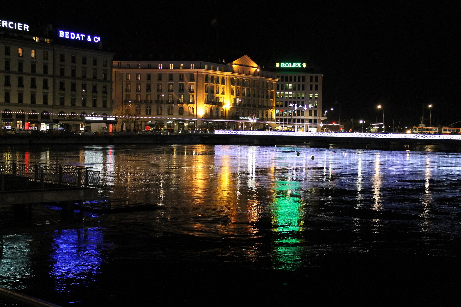 White festive lights line a bridge in Geneva, with illuminated waterfront buildings reflected in the dark river below at night.