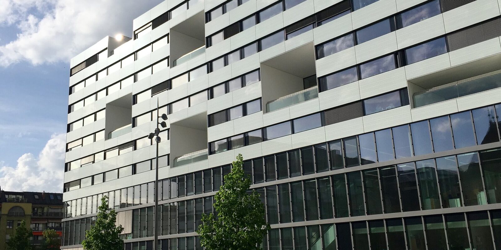 The white Gustave Julliard building at Geneva University Hospitals rises above trees and pedestrians beneath a partly cloudy sky.