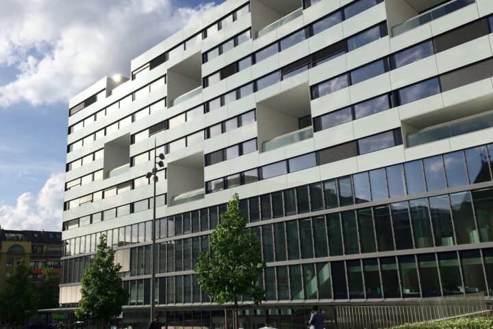 The white Gustave Julliard building at Geneva University Hospitals rises above trees and pedestrians beneath a partly cloudy sky.