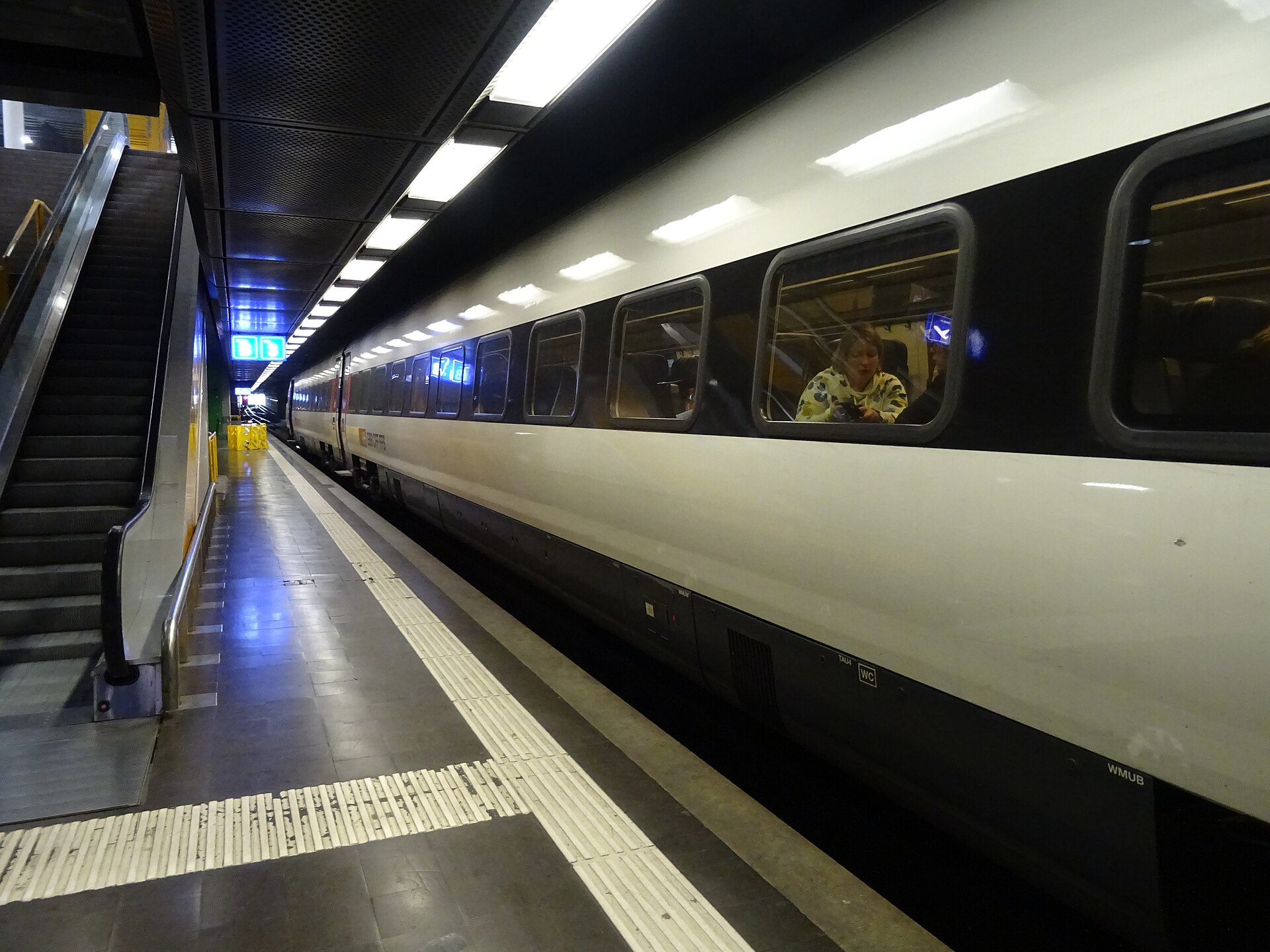 A white SBB train stands beside an escalator on the underground platform at Geneva Airport railway station.