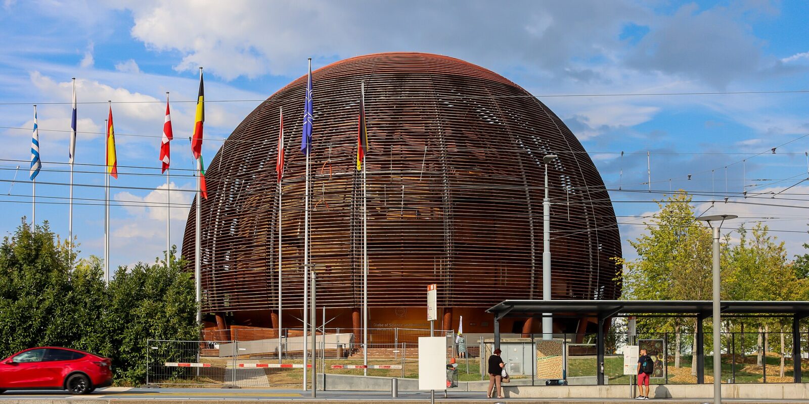 The wooden CERN Globe rises behind flags and a tram platform in Meyrin, with pedestrians waiting beside the road.