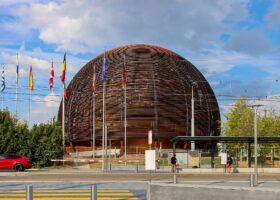 The wooden CERN Globe rises behind flags and a tram platform in Meyrin, with pedestrians waiting beside the road.