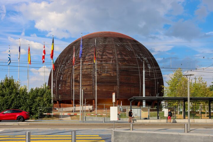 The wooden CERN Globe rises behind flags and a tram platform in Meyrin, with pedestrians waiting beside the road.