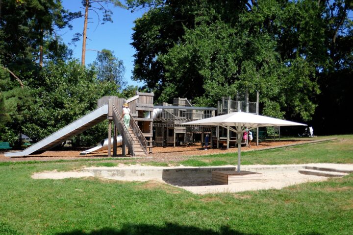 Wooden climbing towers and metal slides stand beneath trees at Parc La Grange in Geneva, beside a sandpit with a sunshade.