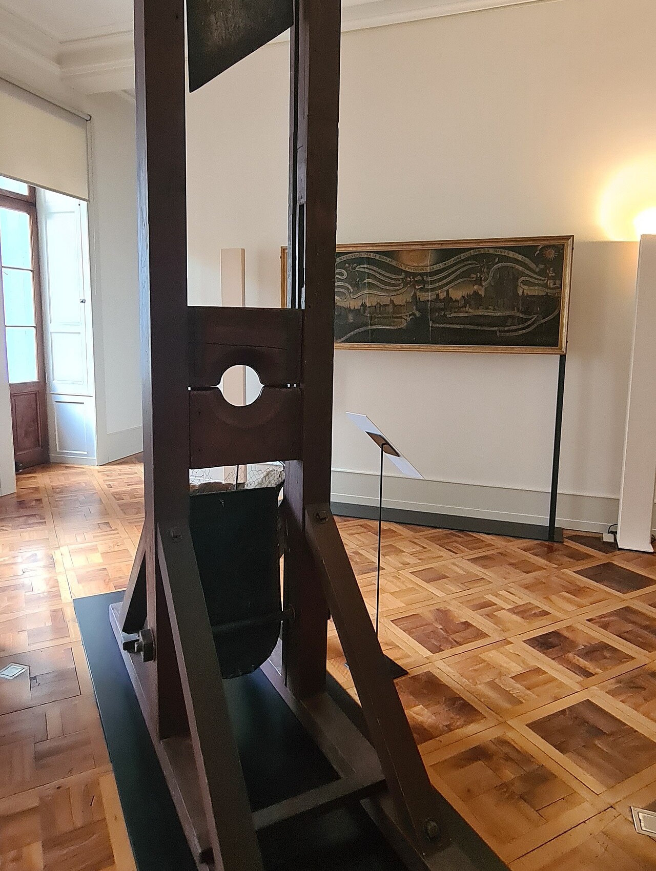 A wooden guillotine reconstruction stands on a low platform in a museum room with a parquet floor, moulded cornice and a framed painting behind it.