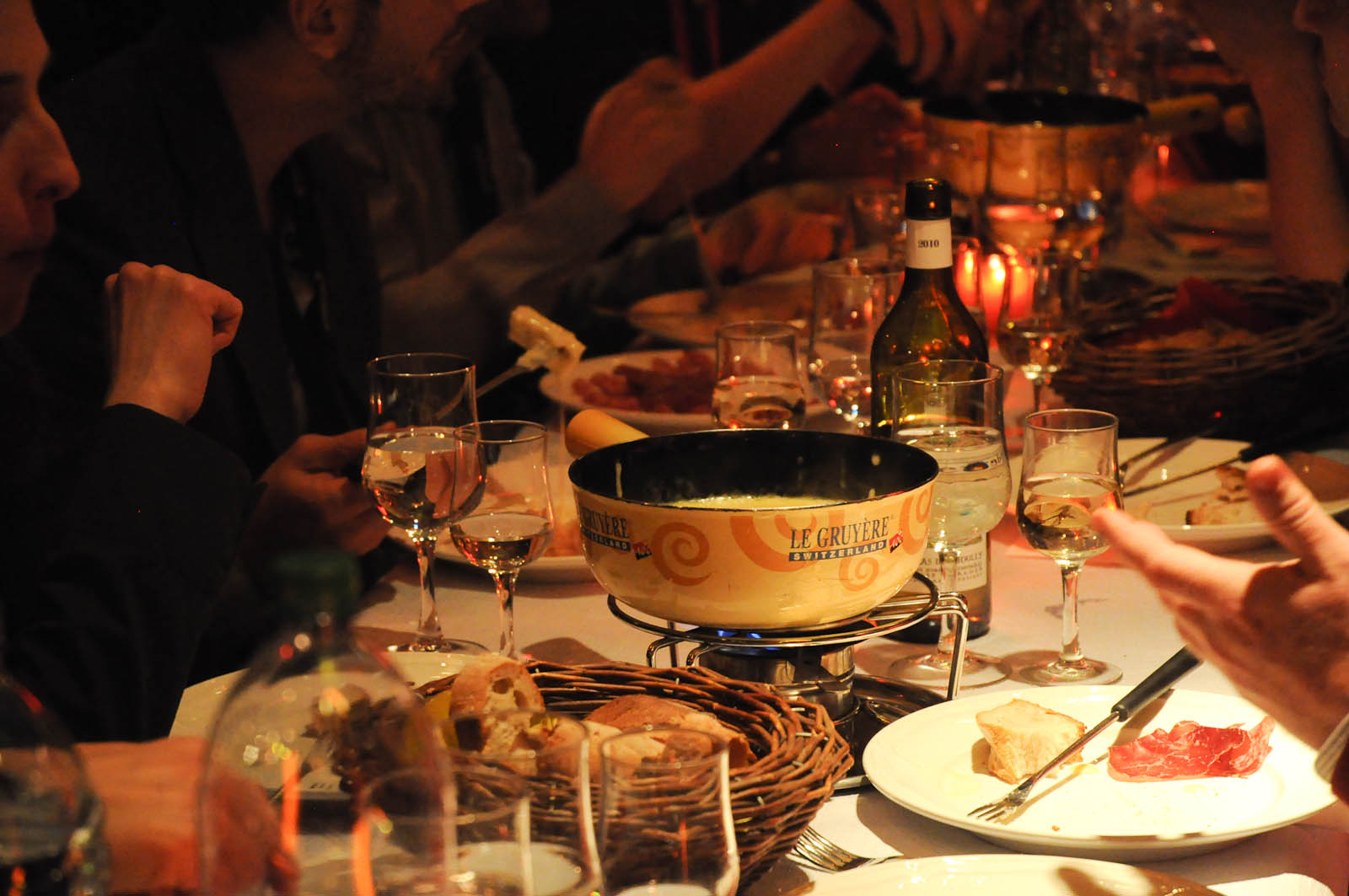 A yellow cheese fondue pot warms over a blue flame among bread baskets and wine glasses at a shared dinner.