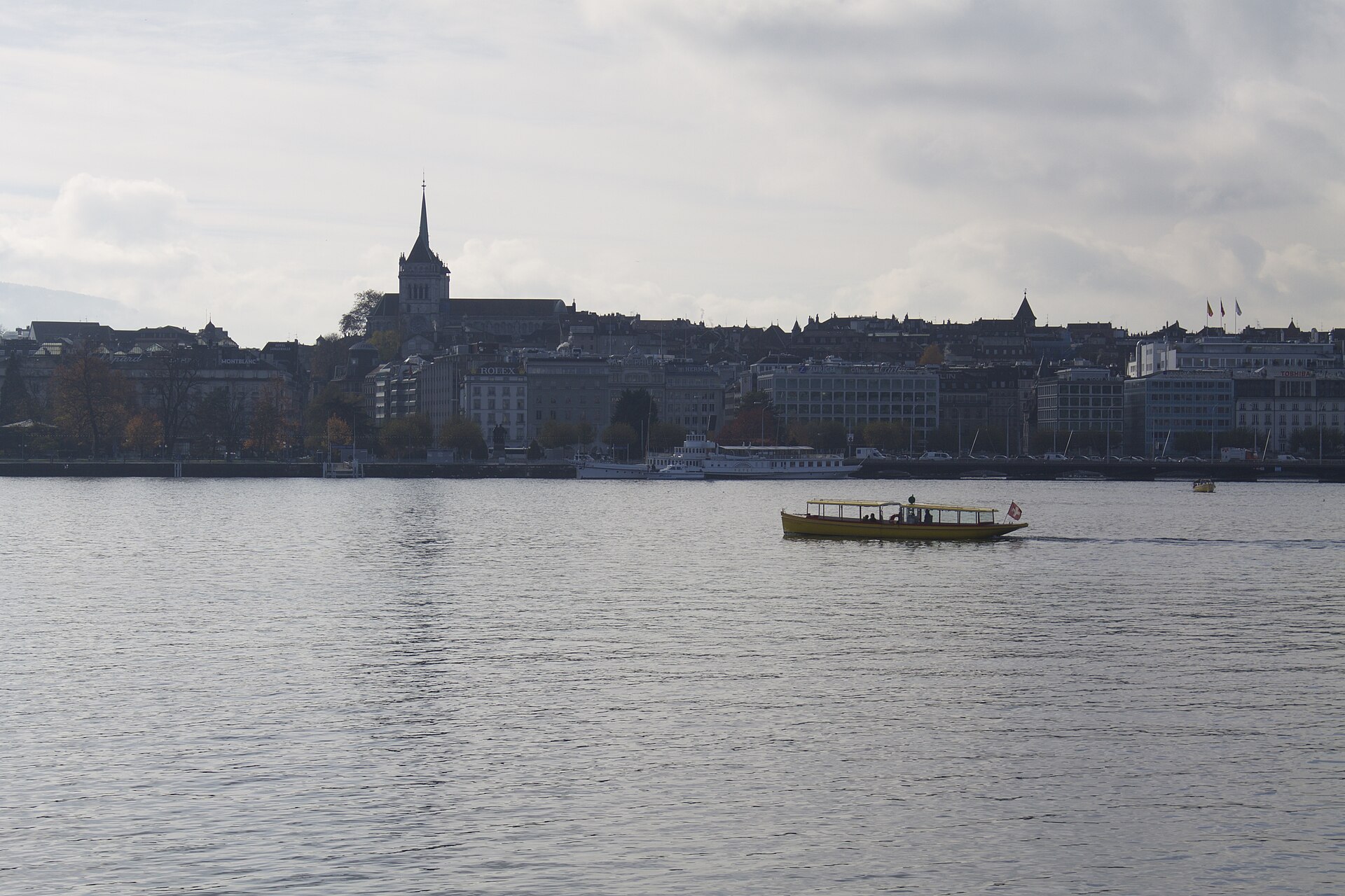 Yellow Mouette water taxi underway across Geneva harbour below the waterfront buildings and towers of Saint Pierre Cathedral.