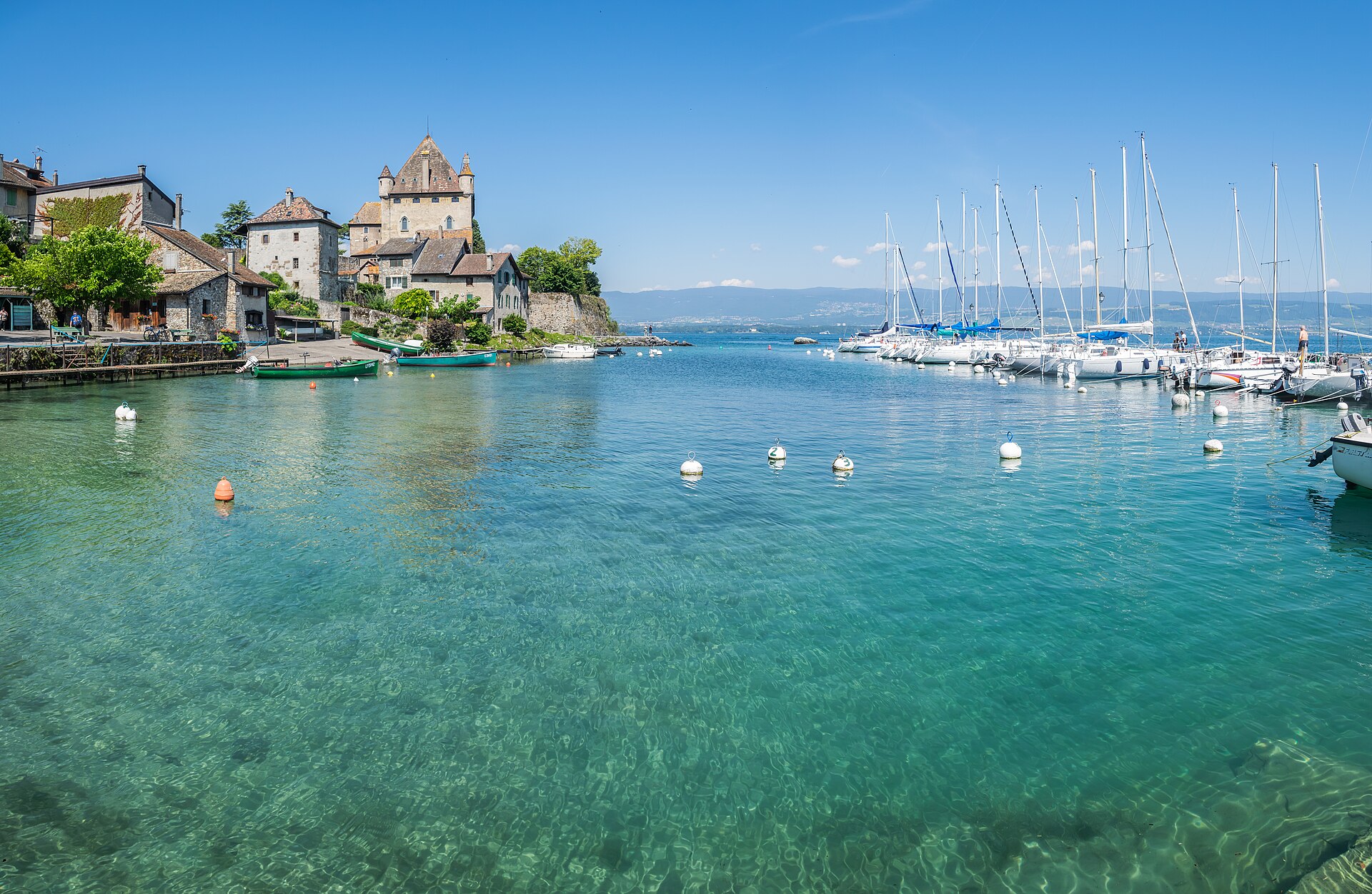 Yvoire castle overlooks clear harbour water, with mooring buoys in the foreground and a line of sailboats beside the village.