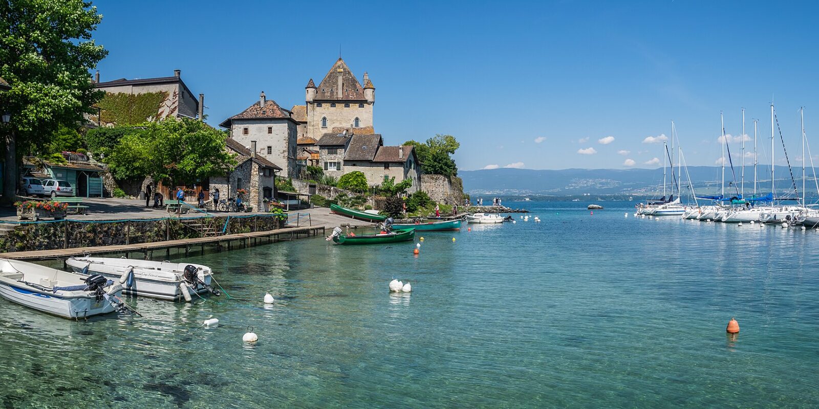 Yvoire's pointed-roof castle overlooks stone houses, small fishing boats and a sailboat harbour on Lake Geneva beneath a clear sky.